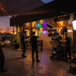 Filmski set House California u večernjoj atmosferi sa produkcijskom ekipom, kamerama i studijskim rasvetnim svetlima u toplim i obojenim tonovima.